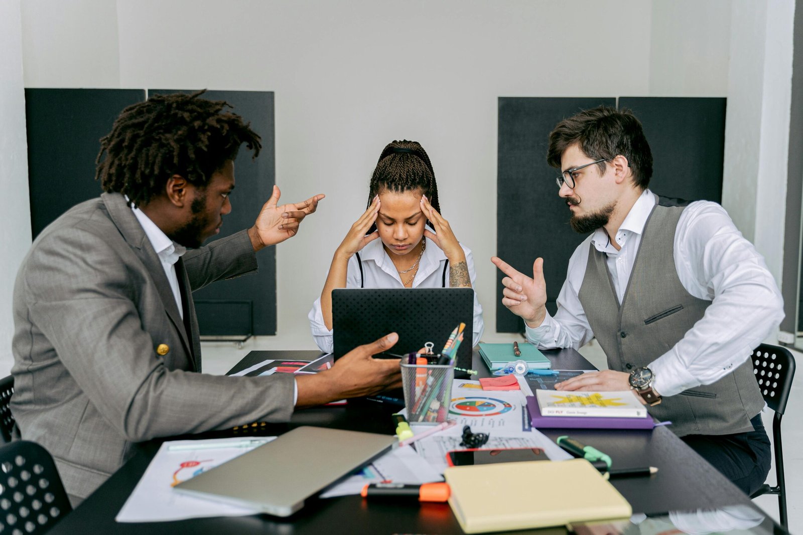Two men argue while a woman looks frustrated at a laptop in an office environment.